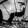 Bodine's Industry" by Schiffer Publishing, Ltd. features a striking black-and-white photo: a boy in a straw hat, holding reins atop old farm equipment, framed by metal spokes against a cloudy sky and open field.