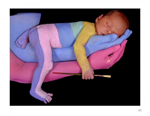 In a scene from Painted Alive by Schiffer Publishing, Ltd., a newborn sleeps on pastel-painted adult hands, its body partly painted and holding a paintbrush—a tender moment of contemporary art and bodypainting set against a black background.