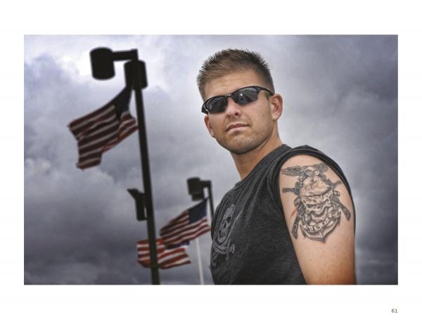 A man stands outdoors in sunglasses and a sleeveless shirt, revealing a large military tattoo on his upper arm, as three American flags wave behind him; featured on the cover of "War Paint" by Schiffer Publishing, Ltd.