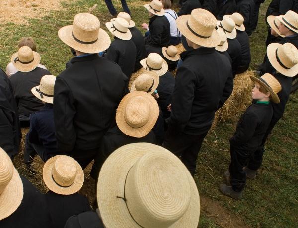 “Faces of Lancaster County” by Schiffer Publishing, Ltd. captures an evocative, artful scene of Amish life—people in black clothing and straw hats gathering outdoors on grass and hay, as seen from above—reflecting Lancaster County’s unique culture.