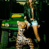 A woman in a black leather jacket and denim shorts stands outdoors with a jaguar on a leash and a green utility vehicle nearby at dusk, evoking the wild spirit of Paradise in Brazil by Schiffer Publishing, Ltd.