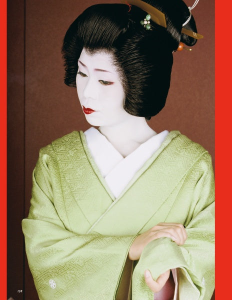 A woman evokes Kyoto geisha as seen in "Geisha & Maiko of Kyoto" by Schiffer Publishing, Ltd., wearing a pale green kimono, white undergarment, white face makeup, red lips, and decorated black hair styled with ornate hairpieces.