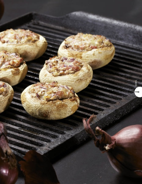 Stuffed mushrooms with a browned filling are grilled to perfection on a ridged pan, ideal for trying new recipes from Pure BBQ! by Schiffer Publishing, Ltd. Whole red onions sit in the foreground on a dark surface.