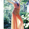 A woman in a peach dress poses outdoors amid greenery and white flowers, her arm raised. She wears sunglasses and a necklace, modeling styles from "Emerging Fashion Designers 3" by Schiffer Publishing, Ltd.