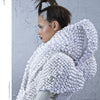 Against a gray, cracked wall, a person with styled brown hair and silver accents models an elaborate white outfit with a sculptural collar—showcasing innovative designs from "Emerging Fashion Designers 3" by Schiffer Publishing, Ltd.