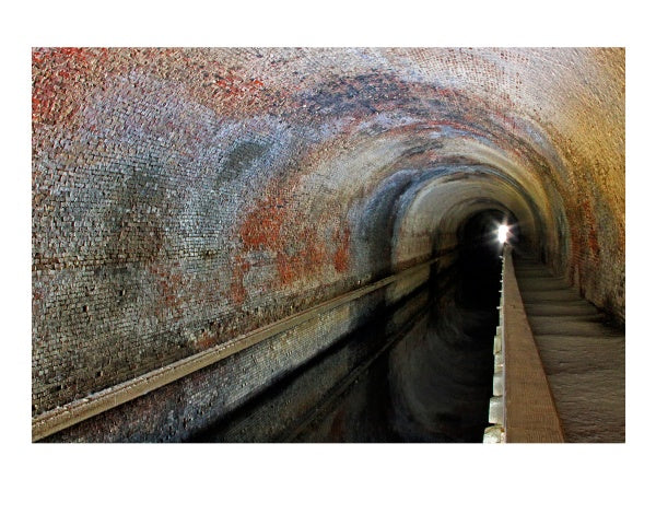 A brick-lined tunnel with a narrow walkway curves alongside a canal, illuminated by light at the end—echoing the historic journey featured in The Chesapeake and Ohio Canal by Schiffer Publishing, Ltd.