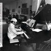 A man in a cap plays piano amid paintings and sculptures in an art-filled studio, as featured in "Jazz in Available Light" by Schiffer Publishing, Ltd.—a book capturing the creative essence of jazz through striking photography.