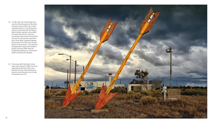 Two large, weathered arrows in overgrown grass beside abandoned buildings under cloudy skies evoke the haunting beauty of America’s ghost towns as seen in Lost Places by Schiffer Publishing, Ltd.