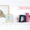 A white shelf displays "The Little Book of Flower Pressing" by Schiffer Publishing, Ltd., surrounded by botanical frames, vases, DIY letters, a green palm leaf, and colorful pennant flags. "THE PROJECTS" is written in red above.